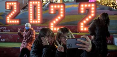 Dos jóvenes posan para tomarse una selfi dando la bienvenida al 2022 en Pekín, China, mientras otra persona saluda al fondo.